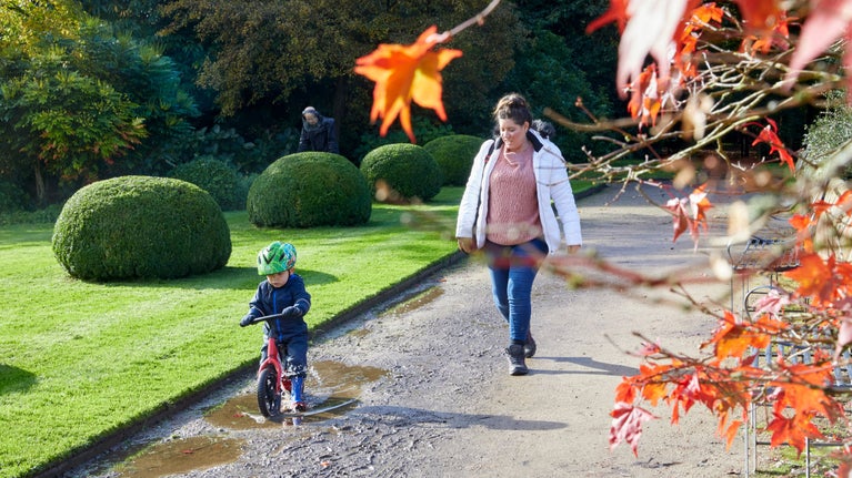 A mother with her toddler, who is on a balance bike going through a puddle, walk along a path through the graden